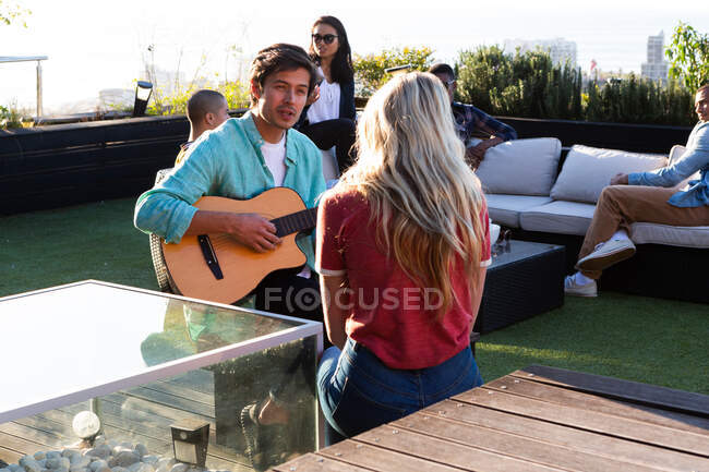 Vista frontale di un uomo caucasico appeso su una terrazza sul tetto in una giornata di sole, che suona la chitarra davanti al suo amico, con la gente che parla sullo sfondo — Foto stock
