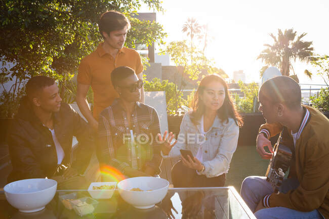 Vista frontale di un gruppo multietnico di amici appesi su una terrazza sul tetto in una giornata di sole, seduti a un tavolo a parlare, uno dei quali con una chitarra in mano — Foto stock