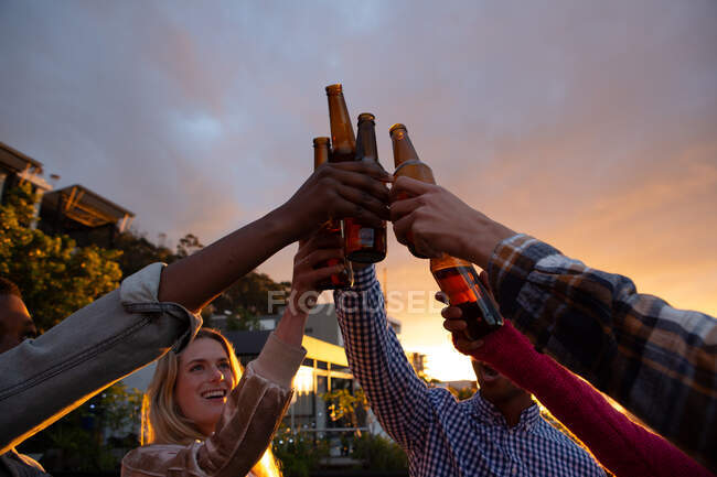 Вид спереду на багатоетнічну групу друзів, які висять на терасі на даху з небом заходу сонця, тримаючи пляшки пива і роблячи тости — стокове фото