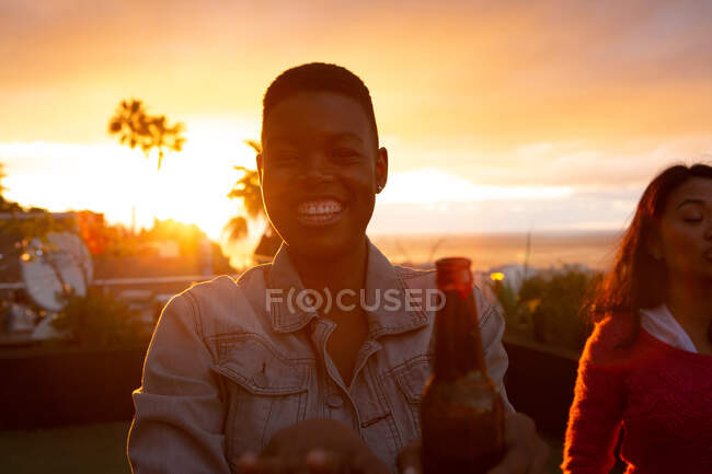 Retrato de un afroamericano colgando en una azotea con un cielo al atardecer, mirando a la cámara y sonriendo, sosteniendo una botella de cerveza, con otra persona en el fondo - foto de stock
