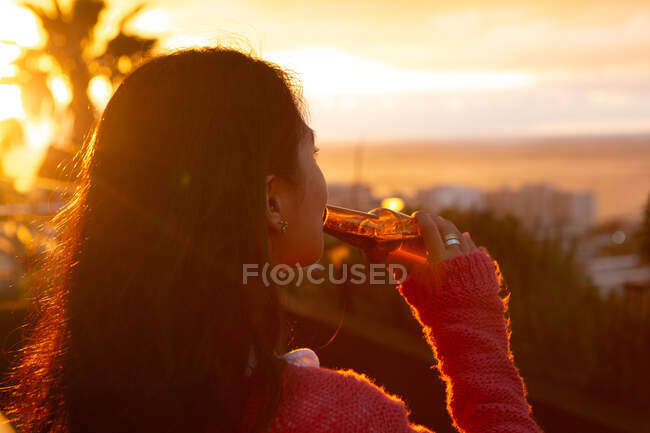 Vue arrière rapprochée d'une femme métisse qui traîne sur une terrasse sur le toit avec un ciel couchant, tenant une bouteille de bière et buvant — Photo de stock