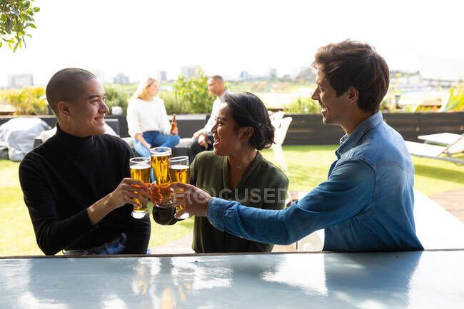 Vista frontal de um grupo multi-étnico de amigos pendurados em um terraço em um dia ensolarado, fazendo um brinde, sorrindo — Fotografia de Stock