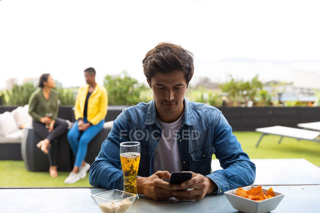 Попереду Кавказький чоловік, який висів на терасі даху в сонячний день, користуючись смартфоном, склянкою пива поруч з ним на столі, і люди говорять на задньому плані. — стокове фото
