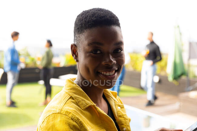 Portrait of an African American woman hanging out on a roof terrace on a sunny day, looking at camera and smiling, with people talking in the background — Stock Photo