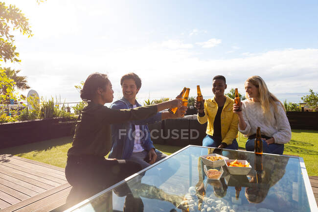 Вид спереду на багатоетнічну групу друзів, які висять на терасі на даху в сонячний день, сидячи за столом, тримаючи пляшки пива, роблячи тост, посміхаючись — стокове фото