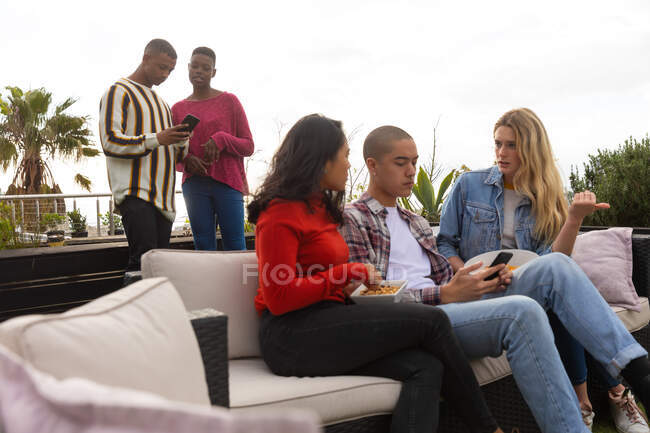 Vista frontale di un gruppo multietnico di amici appesi su una terrazza sul tetto in una giornata di sole, utilizzando uno smartphone e parlando — Foto stock