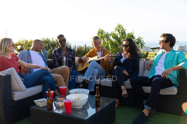 Vista frontale di un gruppo multietnico di amici appesi su una terrazza sul tetto in una giornata di sole, uno di loro che suona la chitarra — Foto stock