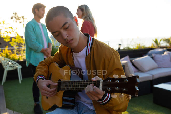 Front view of a mixed race man hanging out on a roof terrace on a sunny day, sitting and playing the guitar, with people talking in the background — Stock Photo