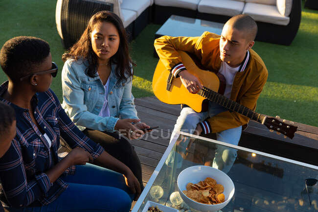 Високий кут зору багатоетнічної групи друзів, що висять на терасі на даху в сонячний день, сидячи за столом і розмовляючи, один з них тримає гітару — стокове фото