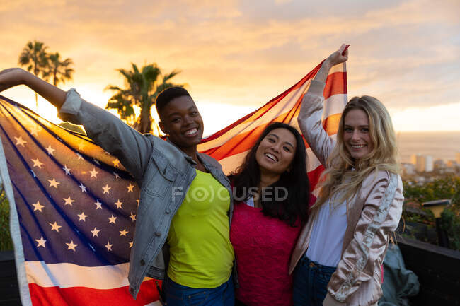 Ritratto di un gruppo multietnico di amici che gironzolano su una terrazza sul tetto con cielo al tramonto, guardano la macchina fotografica e sorridono, sorreggono una bandiera americana — Foto stock