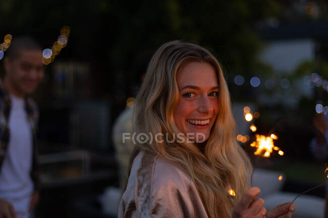Retrato de uma mulher caucasiana pendurada em um terraço, olhando para a câmera e sorrindo, segurando um sparkler, com pessoas no fundo — Fotografia de Stock
