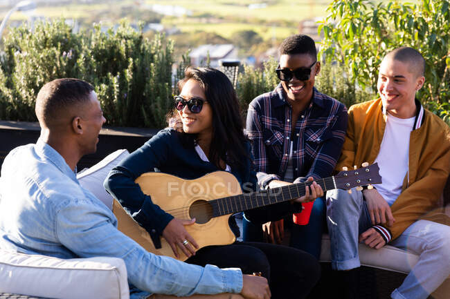 Vista frontal de un grupo multiétnico de amigos pasando el rato en una terraza en un día soleado, uno de ellos tocando la guitarra - foto de stock