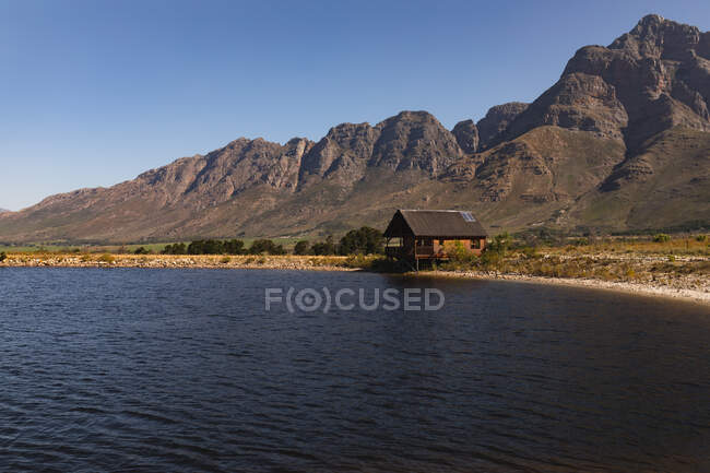 Veduta mozzafiato di una cabina solitaria in legno sulla riva di un lago, vicino alle montagne, in una giornata di sole — Foto stock