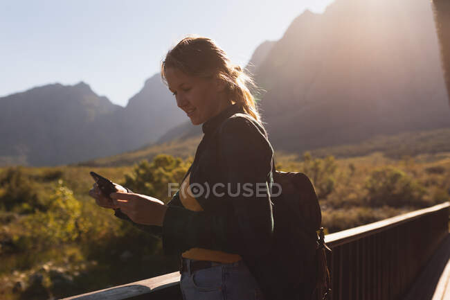 На вигляд кавказька жінка добре проводить час, подорожуючи горами, стоїть на балконі кабіни, користуючись смартфоном, у сонячний день. — стокове фото