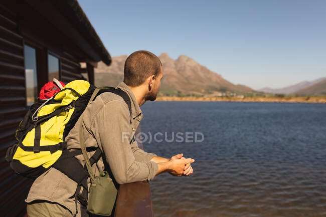 Vista lateral de um homem caucasiano se divertindo em uma viagem às montanhas, de pé em uma varanda de cabine, apreciando sua vista, apoiando-se em uma barreira, em um dia ensolarado — Fotografia de Stock