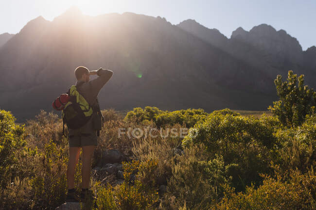 Задний вид кавказца, хорошо проводящего время в поездке в горы, стоящего на поле под горами, наслаждающегося своим видом, защищающего глаза от солнца, в солнечный день — стоковое фото