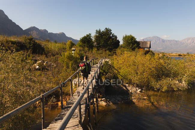 Vista trasera de un hombre caucásico pasándola bien en un viaje a las montañas, de pie sobre un puente, disfrutando de su vista, caminando hacia una cabaña, en un día soleado - foto de stock