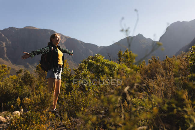 Vista lateral de una mujer caucásica pasándola bien en un viaje a las montañas, de pie en un campo debajo de las montañas, disfrutando de su vista, sosteniendo sus brazos abiertos, en un día soleado - foto de stock
