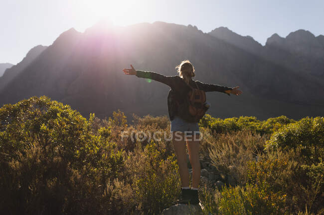 Задний вид на кавказку, хорошо проводящую время в поездке в горы, стоящую на поле под горами, наслаждающуюся своим видом, широко держащую руки, в солнечный день — стоковое фото