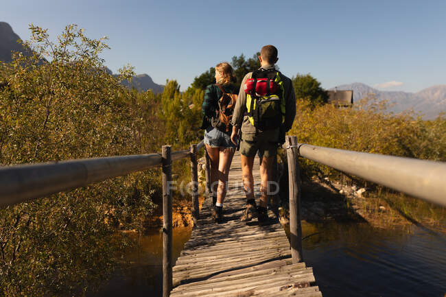 Visão traseira de um casal caucasiano se divertindo em uma viagem às montanhas, andando em uma ponte de madeira, em um dia ensolarado — Fotografia de Stock