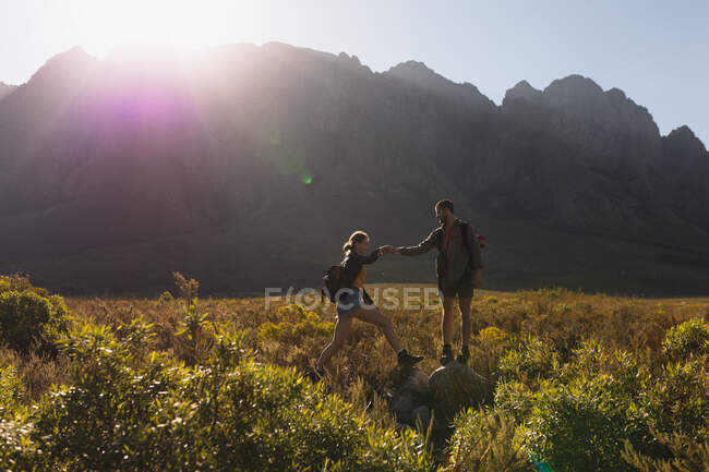 Vista laterale di una coppia caucasica che si diverte durante una gita in montagna, camminando su un campo sotto le montagne, un uomo sta aiutando una donna a salire su una roccia, in una giornata di sole — Foto stock