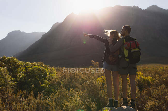 Вид сзади на кавказскую пару, хорошо проводящую время в поездке в горы, гуляющую по полю под горами, стоящую на скале вместе, женщину, указывающую на что-то, в солнечный день — стоковое фото