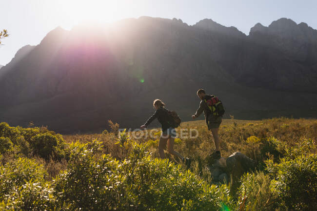 Вид сбоку на кавказскую пару, хорошо проводящую время в поездке в горы, гуляющую по полю под горами, спускающуюся со скалы вместе, в солнечный день — стоковое фото