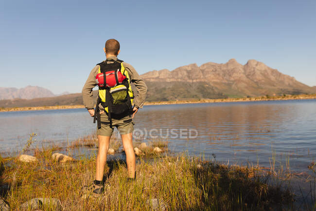 Rückansicht eines kaukasischen Mannes, der sich bei einem Ausflug in die Berge amüsiert, am Ufer steht und die Aussicht genießt, an einem sonnigen Tag — Stockfoto
