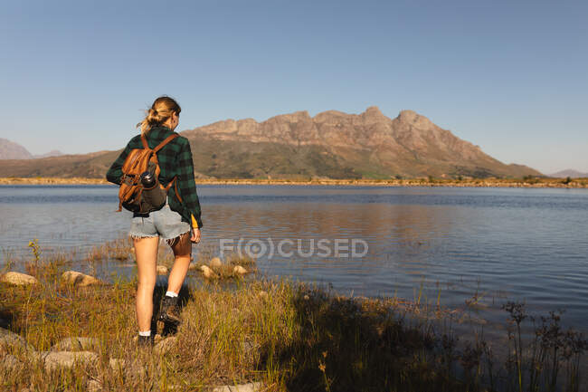 Vista trasera de una mujer caucásica pasándola bien en un viaje a las montañas, de pie en una orilla, disfrutando de su vista, en un día soleado - foto de stock