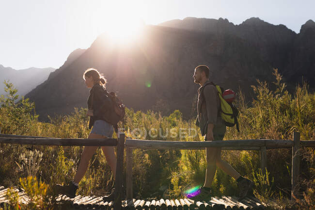 Seitliche Nahaufnahme eines kaukasischen Paares, das an einem sonnigen Tag eine gute Zeit bei einem Ausflug in die Berge hat, auf einer Holzbrücke spazieren geht — Stockfoto