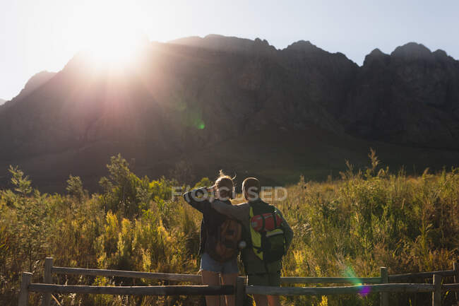 Vista trasera de cerca de una pareja caucásica pasar un buen rato en un viaje a las montañas, de pie en un puente de madera, una mujer está protegiendo sus ojos de la luz del sol, en un día soleado - foto de stock
