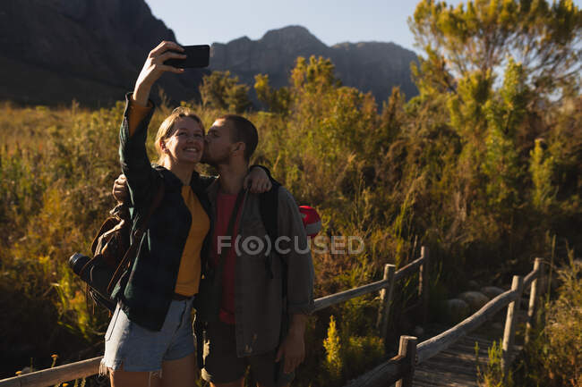 Vista frontal de cerca de una pareja caucásica pasando un buen rato en un viaje a las montañas, de pie en un puente de madera, tomando una selfie, un hombre está besando a su pareja en una mejilla, en un día soleado - foto de stock