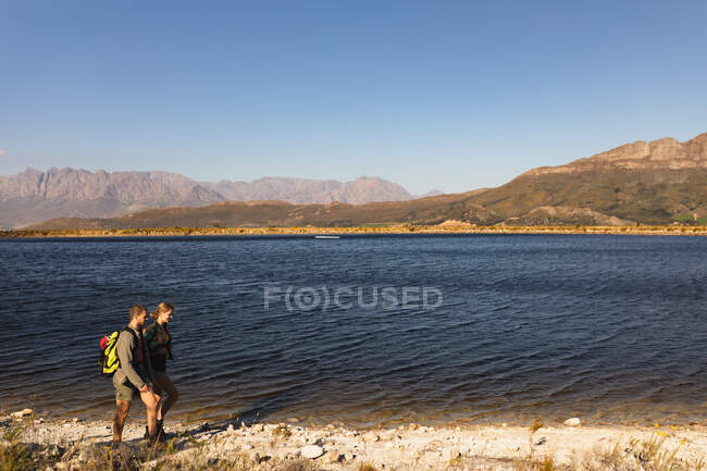 Vista lateral de um casal caucasiano se divertindo em uma viagem às montanhas, andando em um caminho, em uma costa, em um dia ensolarado — Fotografia de Stock