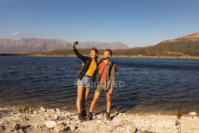 Front view close up of a Caucasian couple having a good time on a trip to the mountains, standing on a path, on a shore, taking a selfie, on a sunny day — Stock Photo