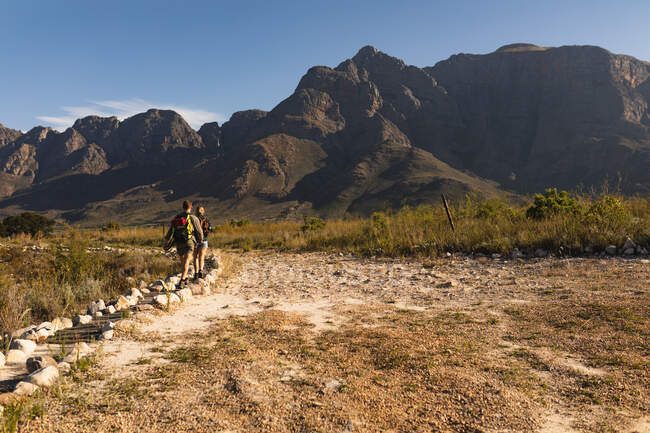 Vue arrière d'un couple caucasien s'amusant lors d'un voyage à la montagne, marchant sur un sentier, par une journée ensoleillée — Photo de stock
