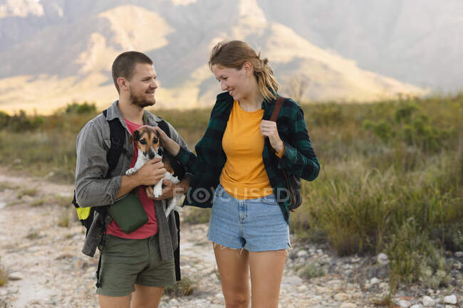 Vue de face gros plan d'un couple caucasien s'amusant lors d'un voyage à la montagne, debout sur un chemin, un homme tient un chiot, une femme le caresse, par une journée ensoleillée — Photo de stock