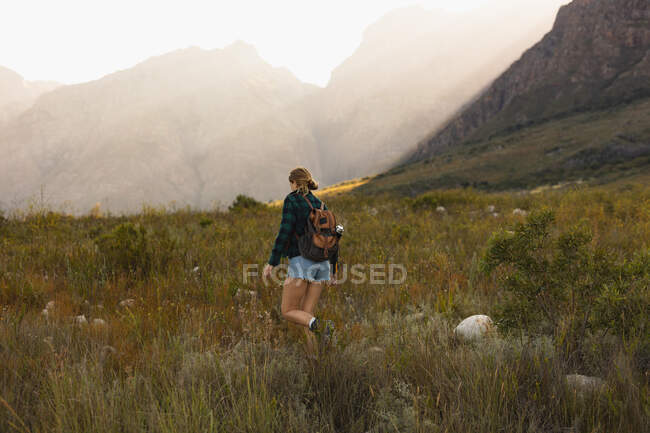 Visão traseira de uma mulher caucasiana se divertindo em uma viagem às montanhas, andando em um campo, em um dia ensolarado — Fotografia de Stock