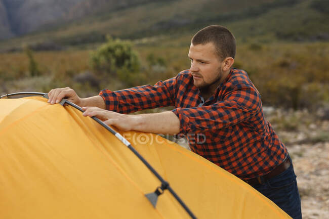 Vista lateral de un hombre caucásico pasándola bien en un viaje a las montañas, levantando una tienda de campaña - foto de stock