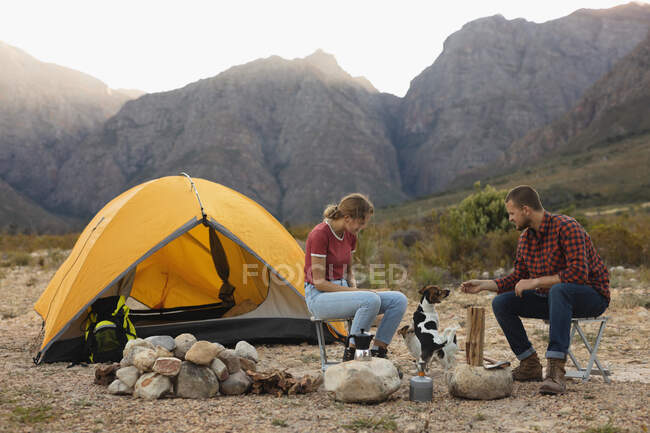 Vista lateral de um casal caucasiano se divertindo em uma viagem às montanhas, sentado perto de uma fogueira, brincando com um cachorro — Fotografia de Stock