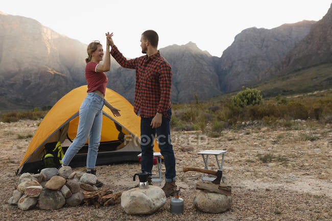 Seitenansicht eines kaukasischen Paares, das eine gute Zeit auf einer Reise in die Berge hat, am Lagerfeuer stehend, High Fiving — Stockfoto