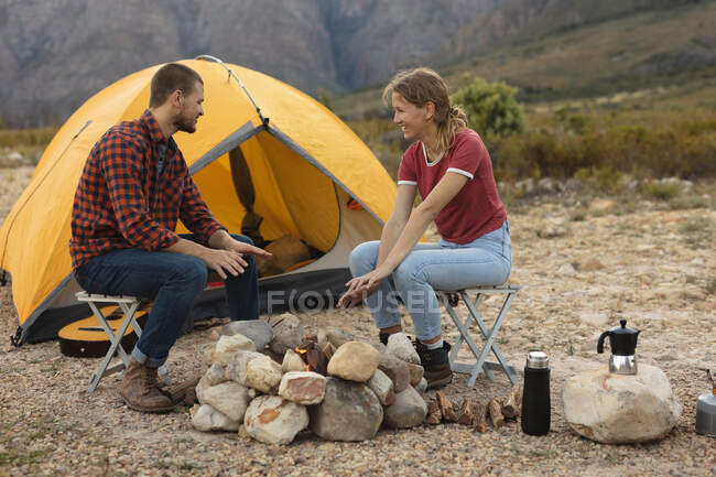 Vista lateral de una pareja caucásica pasándola bien en un viaje a las montañas, sentada junto a una fogata, hablando, sonriendo - foto de stock