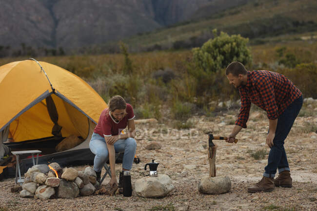 Vista lateral de um casal caucasiano se divertindo em uma viagem às montanhas, um homem está cortando madeira e uma mulher está empilhando — Fotografia de Stock