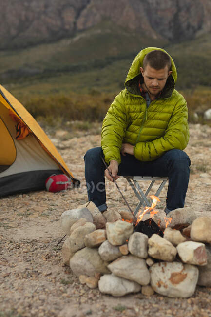 Vue de face d'un homme caucasien s'amusant lors d'un voyage dans les montagnes, assis près d'un feu de camp, regardant le feu — Photo de stock