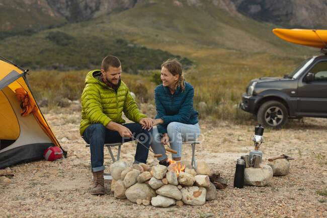 Vista lateral de um casal caucasiano se divertindo em uma viagem às montanhas, sentado perto de uma fogueira, cozinhando salsichas nas varas, de mãos dadas — Fotografia de Stock