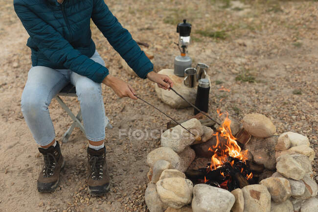 Vue latérale à angle élevé de la femme qui passe un bon moment en voyage à la montagne, assise près d'un feu de camp, faisant cuire des saucisses sur les bâtons — Photo de stock