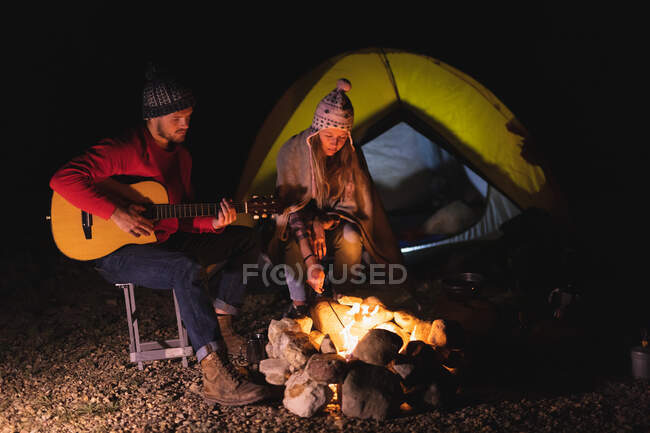 Vista lateral de um casal caucasiano se divertindo em uma viagem para as montanhas, sentado perto de uma fogueira, um homem está tocando uma guitarra, e uma mulher está jogando paus para o fogo — Fotografia de Stock