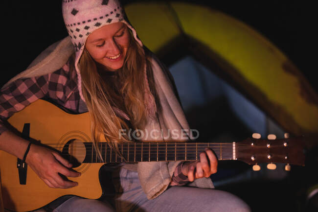Vorderansicht einer kaukasischen Frau, die eine gute Zeit auf einer Reise in die Berge hat, am Lagerfeuer sitzt, Gitarre spielt, lächelt — Stockfoto