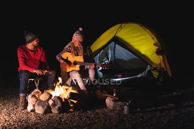 Vista lateral de una pareja caucásica pasándola bien en un viaje a las montañas, sentada junto a una fogata, una mujer está tocando una guitarra, mirándose y sonriendo - foto de stock