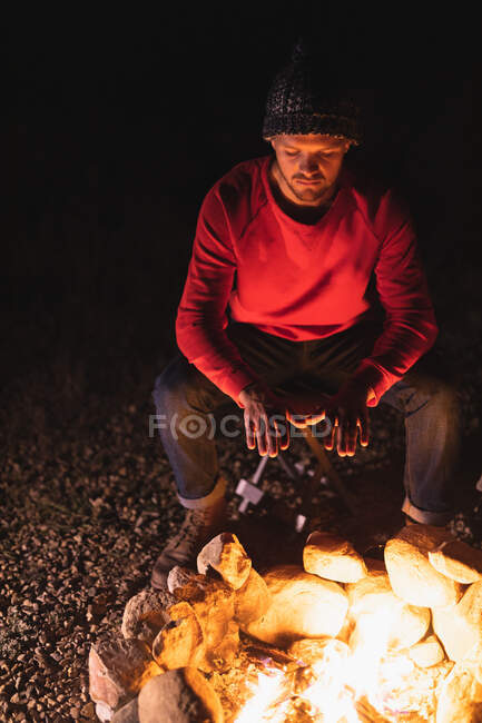 Vue de face d'un homme caucasien s'amusant lors d'un voyage dans les montagnes, assis près d'un feu de camp, se réchauffant les mains — Photo de stock