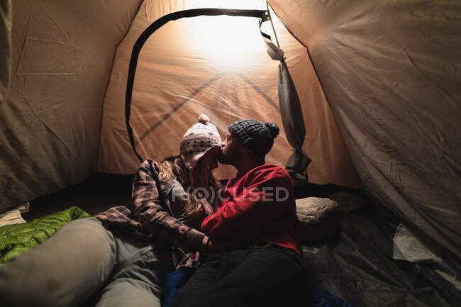 Vista frontal de una pareja caucásica pasando un buen rato en un viaje a las montañas, acostado en una tienda de campaña, un hombre está besando a una mujer en una frente - foto de stock
